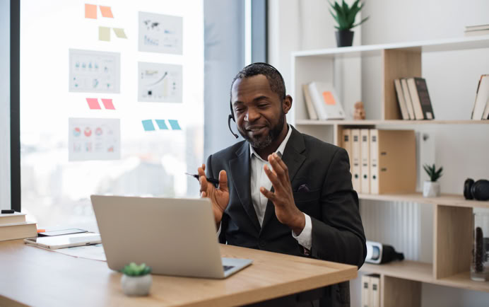 Man in a suit sitting at a desk with a laptop, wearing a headset, gesturing with hands, with shelves and a window in the background.