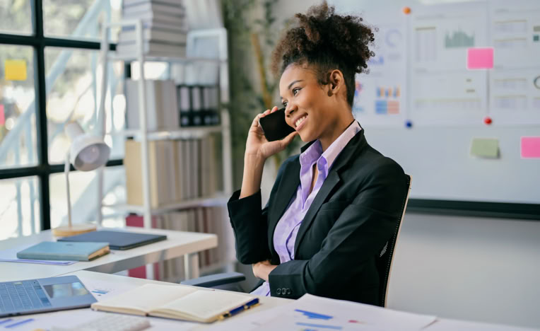 A person in a suit is sitting at a desk, talking on a phone, with documents and a laptop in front of them. A whiteboard with charts and sticky notes is visible in the background.