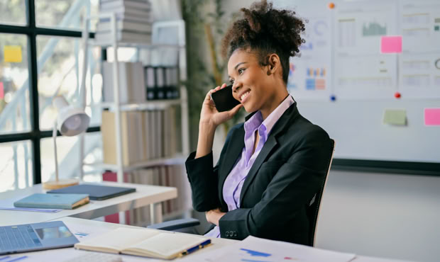 A person in a suit is sitting at a desk, talking on a phone, with documents and a laptop in front of them. A whiteboard with charts and sticky notes is visible in the background.