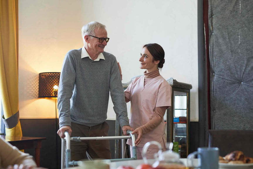 An elderly man using a walker is assisted by a woman in a pink uniform, standing in a room with a table and a lamp.