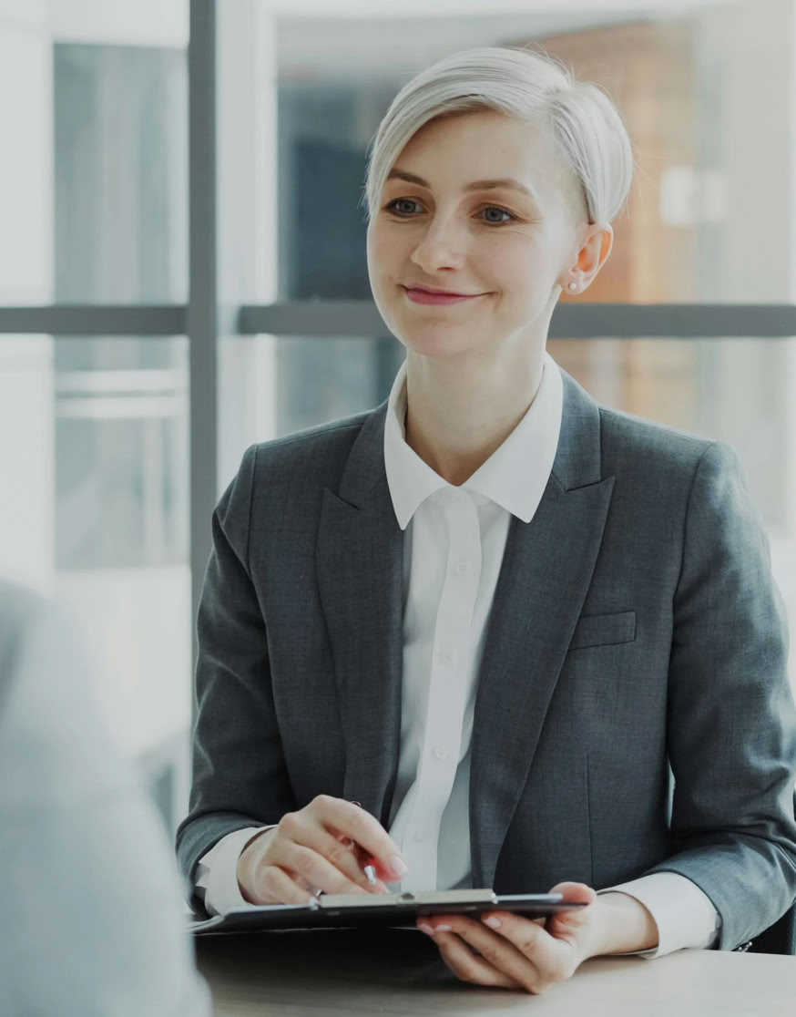 A person with short hair in a suit holding a clipboard, sitting across from another person in an office setting.