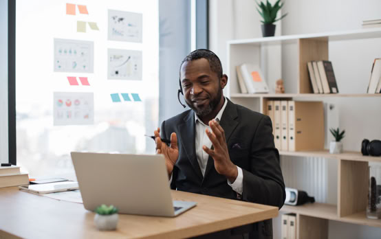Man in a suit sitting at a desk with a laptop, wearing a headset, gesturing with hands, with shelves and a window in the background.