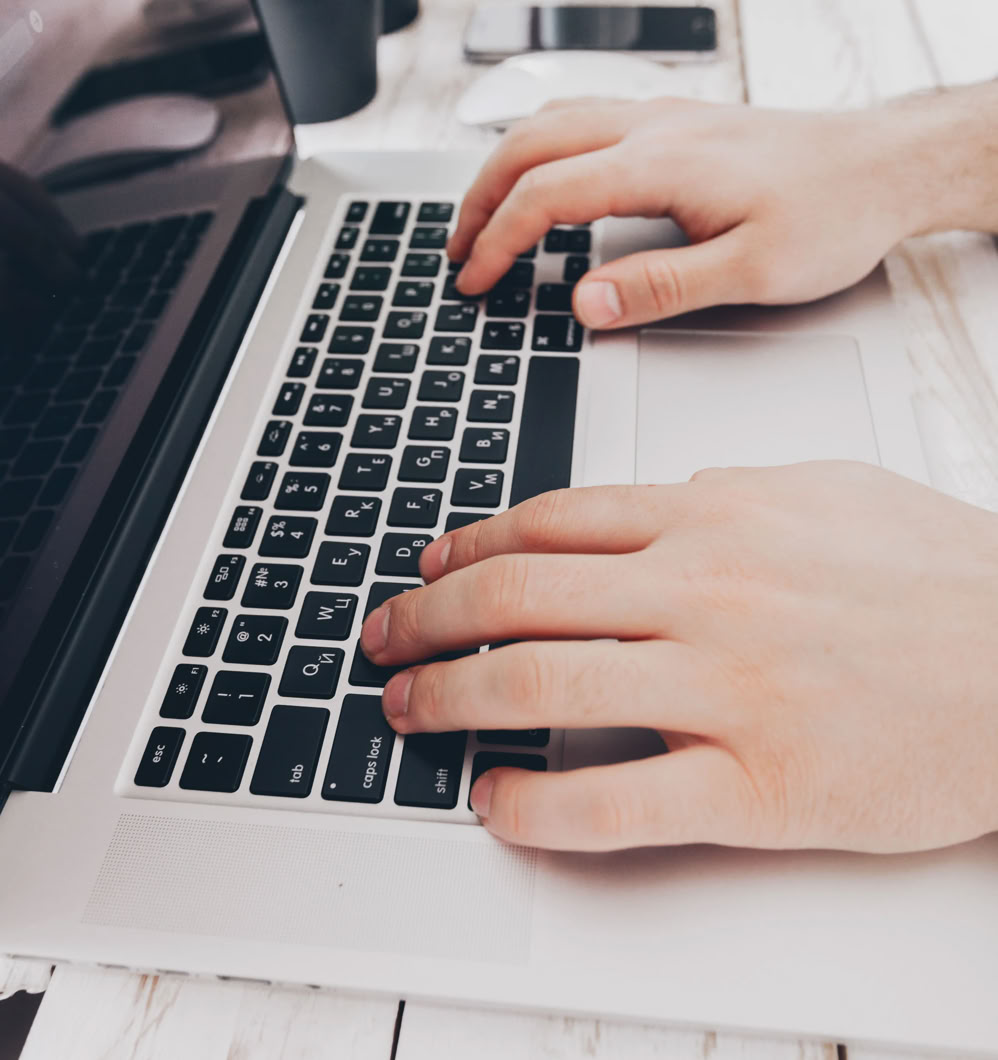 Hands typing on a laptop keyboard on a white desk with a plant, smartphone, and cup in the background.
